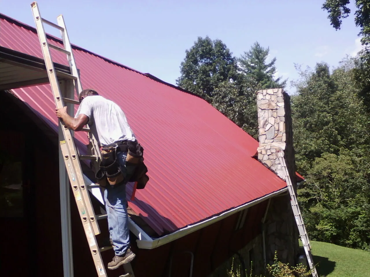 Roofer on a ladder beside a red metal roof and stone chimney — Soffit & Fascia Repair by Roofing Brothers in Slater